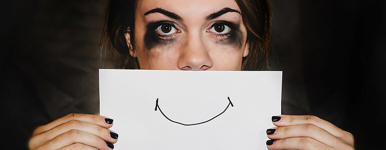 Eine Frau mit dunklen Haaren und schwarz verschmierter Schminke unter den Augen hält ein Blatt Papier mit einem lächelnden Mund vor ihr Gesicht.