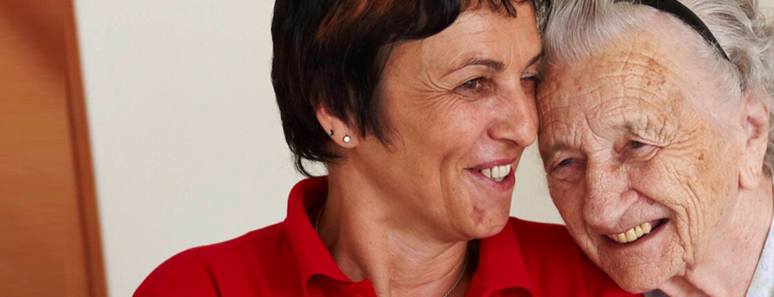 Eine alte Frau lehnt sich an die Schulter einer Pflegerin der Caritas.