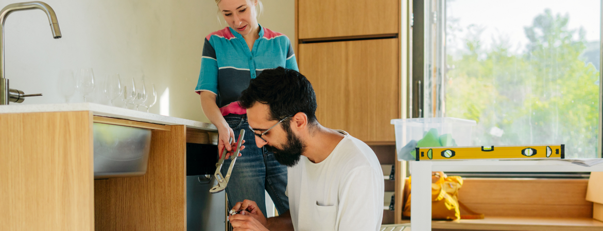 Ein Mann kniet vor einer Spüle, eine Frau steht daneben und reicht ihm eine Zange