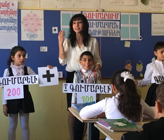 Eine Lehrerin hält in einem Klassenzimmer Unterricht. Neben ihr stehen Schülerinnen, die Schilder mit armenischen Wörtern halten..