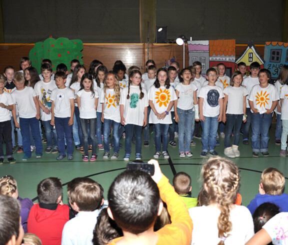 Kinder performen vor Kindern in einem Turnsaal. Die Performer haben alle ein weißes T-Shirt an. 