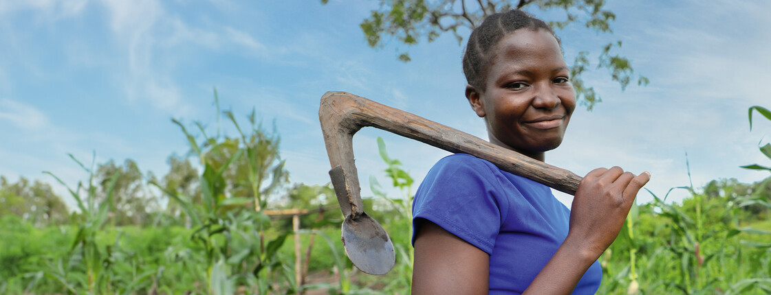 © Nyokabi Kahura_fairpicture Eine junge Frau steht auf einem grünen Feld und trägt eine Hacke über ihrer Schulter. Sie lächelt in die Kamera. Das Bild steht im Kontext der Hungerkampagne 2024.