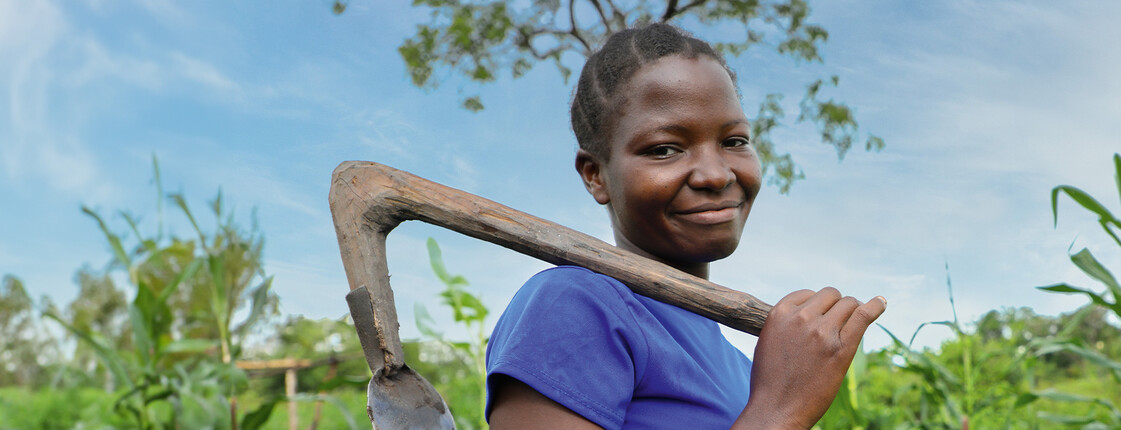 Eine junge Frau steht auf einem grünen Feld und trägt eine Hacke über ihrer Schulter. Sie lächelt in die Kamera. Das Bild steht im Kontext der Hungerkampagne 2024.