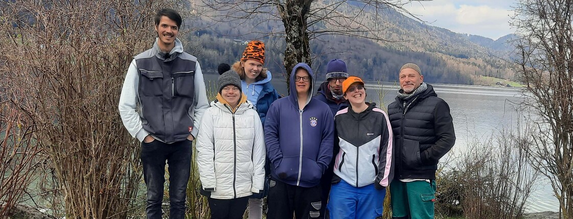 Eine Gruppe von Menschen mit Beeinträchtigung stehen vor einem See und blicken in die Kamera.