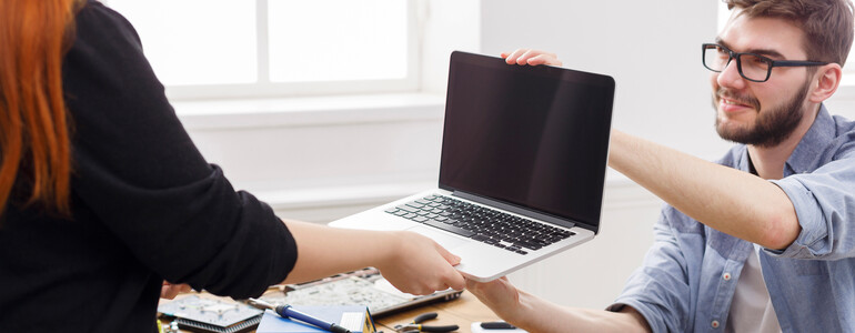 Ein IT-Mitarbeiter, der eine Brille und ein Hemd trägt, übergibt einen aufgeklappten Laptop an eine Frau mit roten Haaren.