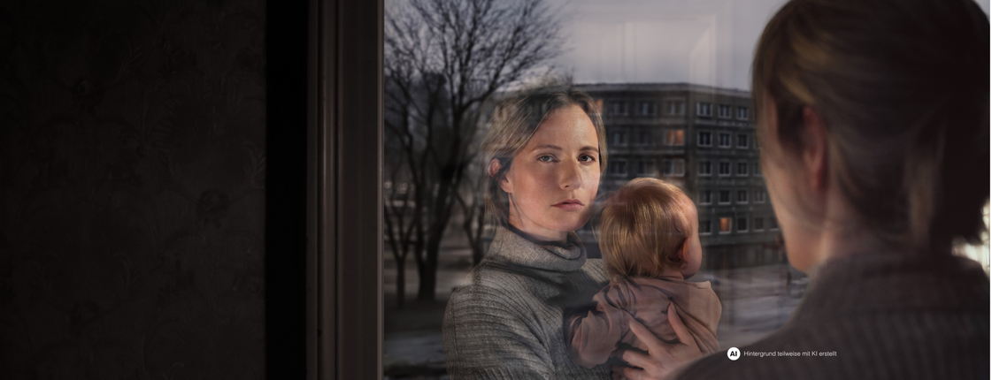 Eine Frau mit Kind am Arm schaut aus dem Fenster. Ihr Gesicht spiegelt sich im Fensterglas wider. Vor dem Fenster ist eine Wohngegend erkennbar.