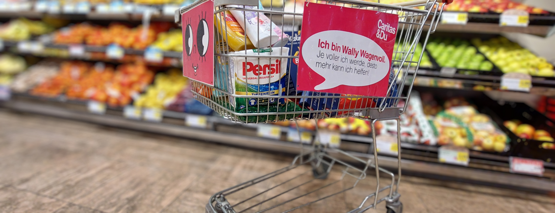im Supermarkt Einkaufswagen mit Caritas-Plakat „Ich bin Wally Wagenvoll“ steht gefüllt mit Produkten vor Obstregal im Supermarkt
