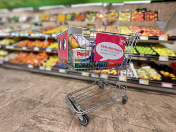 im Supermarkt Einkaufswagen mit Caritas-Plakat „Ich bin Wally Wagenvoll“ steht gefüllt mit Produkten vor Obstregal im Supermarkt