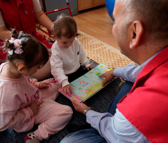Betreuungsperson zeigt zwei Kindern ein buntes Sprachbuch auf dem Boden.