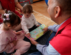 Betreuungsperson zeigt zwei Kindern ein buntes Sprachbuch auf dem Boden.