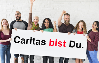 Eine Gruppe von Menschen stehen nebeneinander und halten ein Schild mit der Aufschrift "Caritas bist Du." in den Händen.