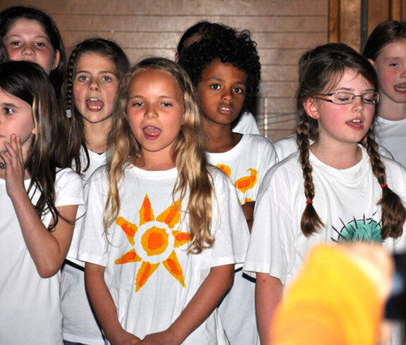 Kinder im Volksschulalter stehen in einem Raum und singen. Sie tragen alle weiße T-Shirts und lange, meist zusammengebundene, Haare. 
