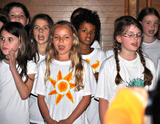 Kinder im Volksschulalter stehen in einem Raum und singen. Sie tragen alle weiße T-Shirts und lange, meist zusammengebundene, Haare. 