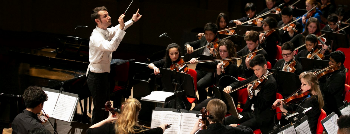 Ein Dirigent steht vor einem Streich-Orchester und dirigiert.