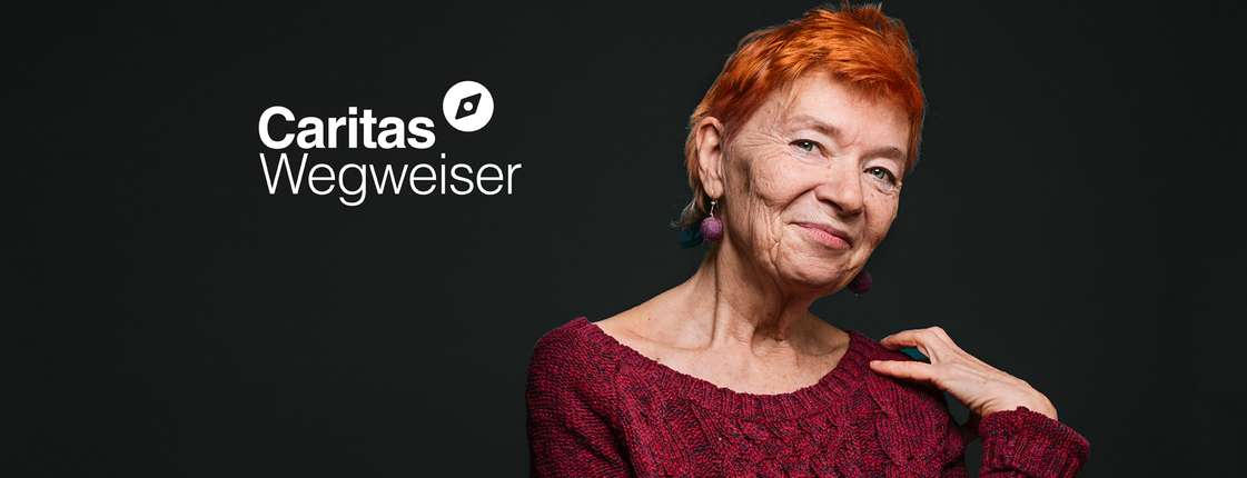 Das Porträt einer Frau ist zu sehen auf schwarzem Hintergrund. Es ist eine ältere Dame die lächelt, sie hat einen rothaarigen Kurzhaarschnitt. Links oben das Logo vom Caritas Wegweiser.