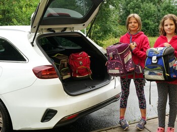 Drei Kinder stehen neben einem offenen Kofferraum des Caritas Autos und halten jeweils eine Schultasche in den Händen 