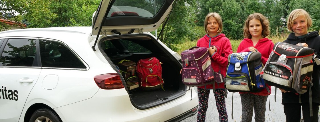 Drei Kinder stehen neben einem offenen Kofferraum des Caritas Autos und halten jeweils eine Schultasche in den Händen 