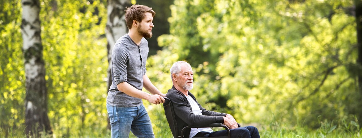 In einem Wald bei schönem Wetter machen ein junger Mann und ein älteren Mann, der im Rollstuhl sitzt, einen Spaziergang.