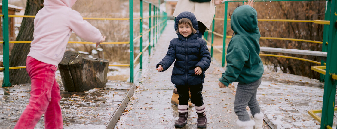 Drei Kinder, die Regenkleidung tragen, spielen vor einem Zentrum im Regen mit ihrer Betreuerin.