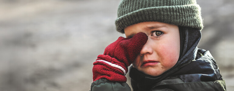 Ein Junge mit einer Schultasche aus Karton steht mitten im Schutt einer Stadt.