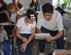 Eine Schülerin mit Behinderungen sitzt mit anderen Kindern in einem Klassenzimmer und erhält von einem älteren Schüler individuelle Unterstützung beim Unterricht.