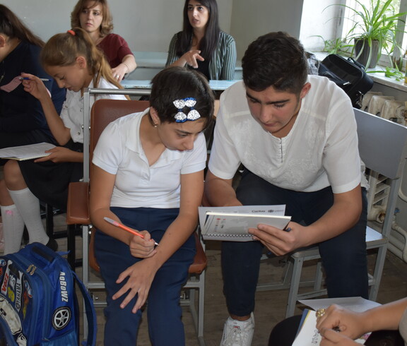 Eine Schülerin mit Behinderungen sitzt mit anderen Kindern in einem Klassenzimmer und erhält von einem älteren Schüler individuelle Unterstützung beim Unterricht.