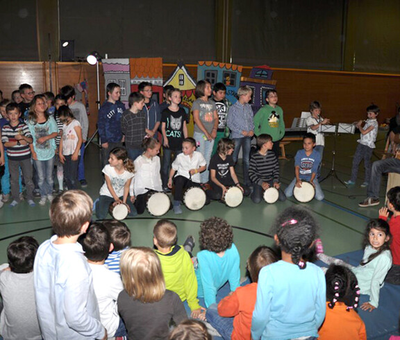 In einem Turnsaal einer Schule sind etliche Kinder versammelt. Kinder spielen Kindern Musik vor. Der Raum ist recht dunkel. Manche stehen, andere sitzen. 