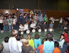 In einem Turnsaal einer Schule sind etliche Kinder versammelt. Kinder spielen Kindern Musik vor. Der Raum ist recht dunkel. Manche stehen, andere sitzen. 