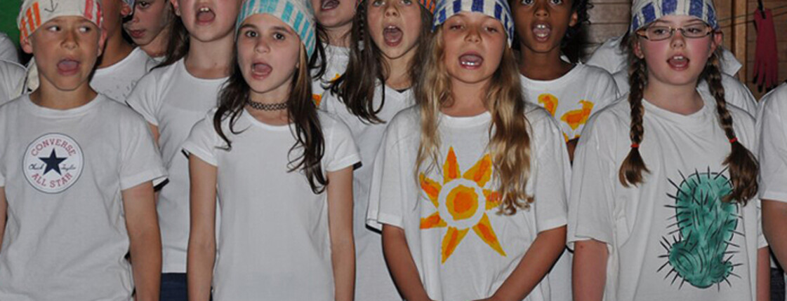 Um die zehn Mädchen im Volksschulalter stehen zusammen und singen. Sie tragen alle lange Haare, weiße T-Shirts und selbstgebastelte Kopfbedekungen. 