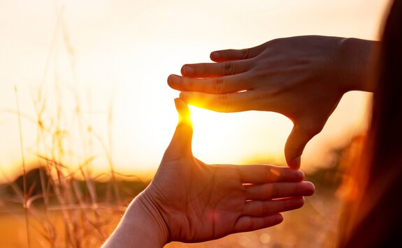 Hände rahmen die untergehende Sonne ein, im Hintergrund verschwommene Gräser und Felder.
