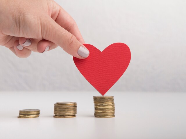 Eine Hand hält ein rotes Papierherz über einen Stapel Münzen