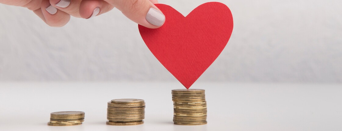 Eine Hand hält ein rotes Papierherz über einen Stapel Münzen