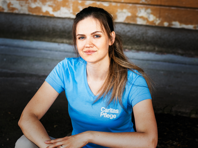 Eine junge Frau mit braunen langen Haaren und Pferdeschwanz sitzt auf einer Stiege, sie trägt ein hellblaues T-Shirt mit Caritas Logo und lächelt in die Kamera.