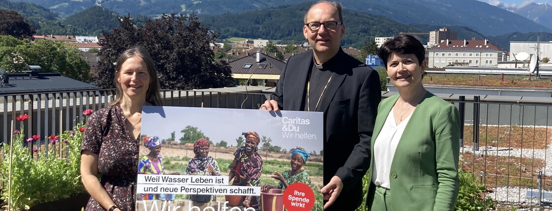 Drei Personen stehen auf einem begrünten Dach und präsentieren ein Plakat der Caritas-Auslandshilfe. 