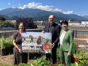 Drei Personen stehen auf einem begrünten Dach und präsentieren ein Plakat der Caritas-Auslandshilfe. 