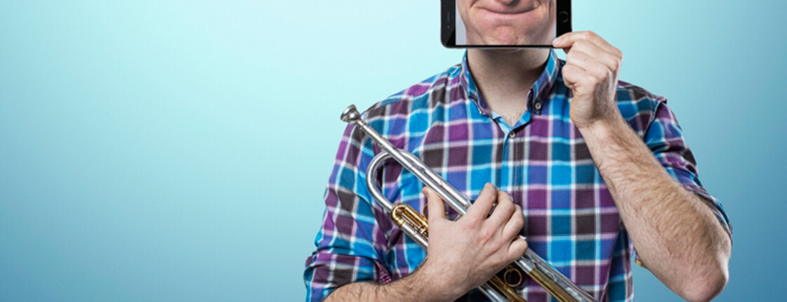 Ein junger Musiker hält eine Trompete in der Hand. Er hält ein Handy vor sein Gesicht, wobei man sein Gesicht durch das Handy sieht. 