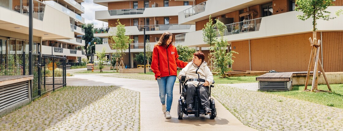 Eine Caritas Mitarbeiterin und eine Frau im Rollstuhl gehen spazieren
