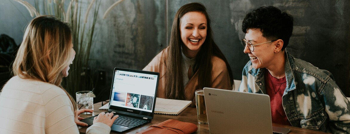 Drei Frauen sitzen an einem Holztisch vor ihren Laptops und lachen zusammen.