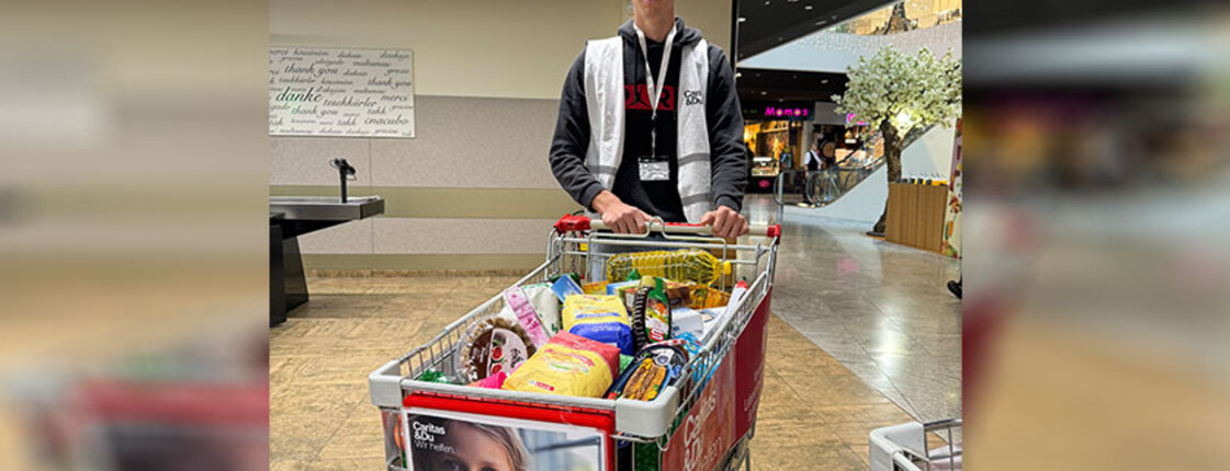 Ein junger Mann steht hinter einem vollen Einkaufswagen und blickt in die Kamera.