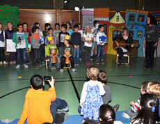 Kinder singen Kindern in einem Turnsaal einer Schule vor. Eine Erwachsene Personen begleitet die Sänger. Der Boden der Halle ist grün. Manche sitzen, mache stehen. 