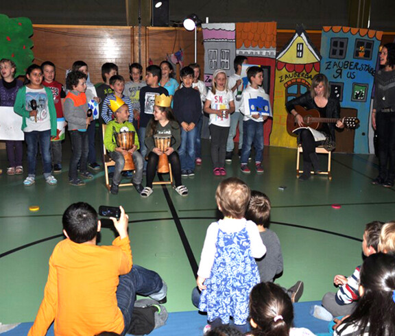 Kinder singen Kindern in einem Turnsaal einer Schule vor. Eine Erwachsene Personen begleitet die Sänger. Der Boden der Halle ist grün. Manche sitzen, mache stehen. 