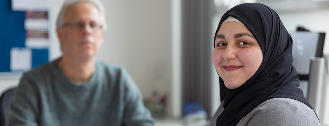 Portrait einer jungen Frau mit schwarzem Kopftuch während eines Beratungsgesprächs. Im Hintergrund sitzt ein Caritas Mitarbeiter an einem Schreibtisch.