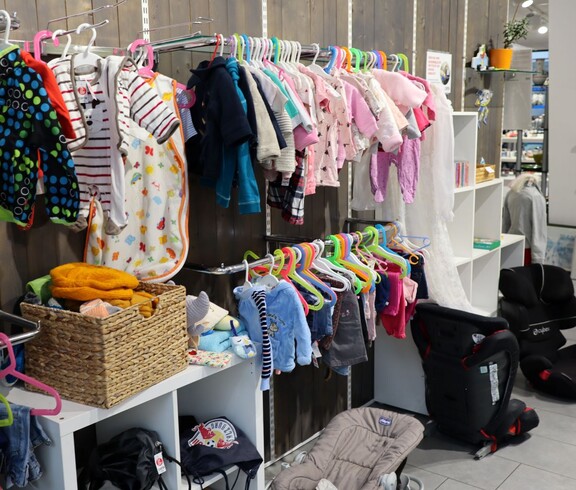 Eine Kleiderstange voller Babykleidung sowie zwei Kindersitzen für das Auto.