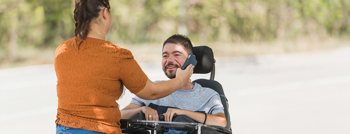  Eine Frau hält einem lächelnden Mann im Rollstuhl ein Smartphone ans Ohr. Sie befinden sich draußen vor einem unscharfen grünen Hintergrund.