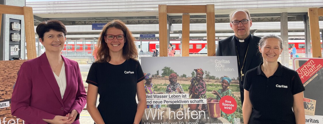 Vier Personen stehen auf einem Bahnsteig und präsentieren Plakate zur Caritas Spendenaktion "Gegen den Hunger"
