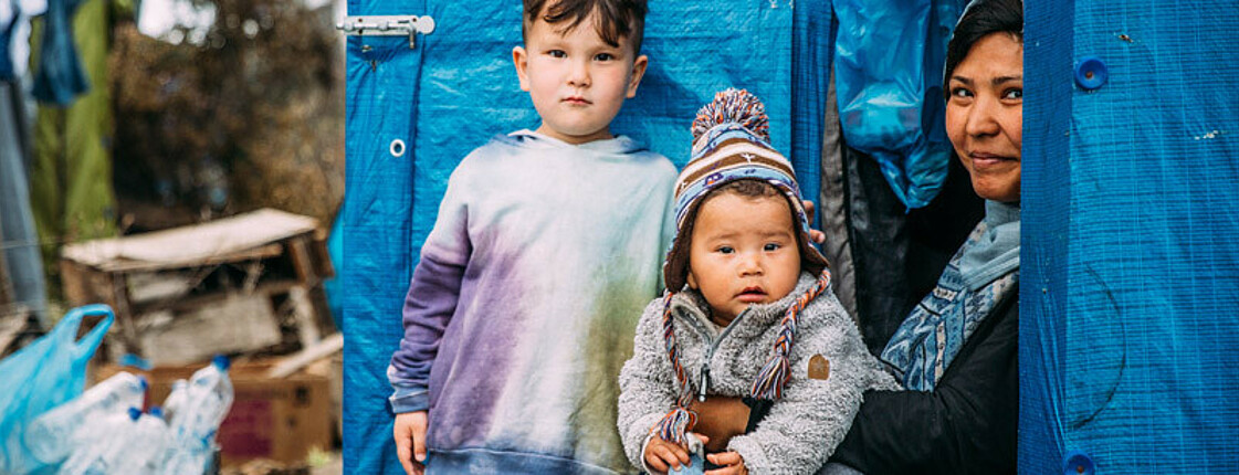 Eine Frau  mit einem Kleinkind im Arm und ein Bub stehen in einem Flüchtlingslager in Griechenland am Eingang ihres Zeltes.