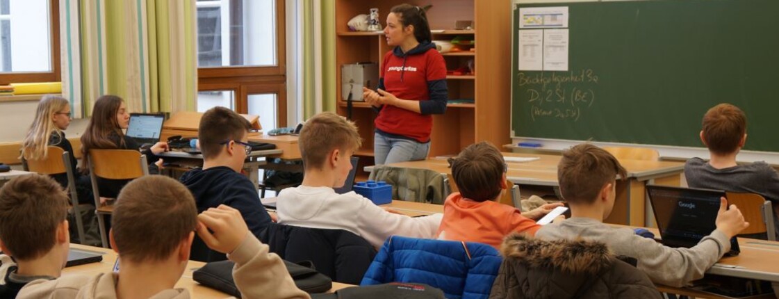 Schüler sitzen in der Klasse. Vorne an der Tafel steht eine Caritas Mitarbeiterin