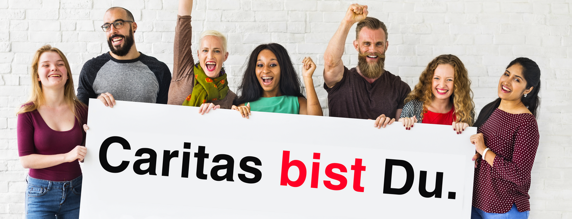 Eine Gruppe von Menschen stehen nebeneinander und halten ein Schild mit der Aufschrift "Caritas bist Du." in den Händen.