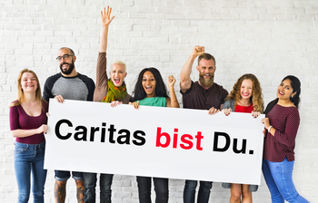 Eine Gruppe von Menschen stehen nebeneinander und halten ein Schild mit der Aufschrift "Caritas bist Du." in den Händen.