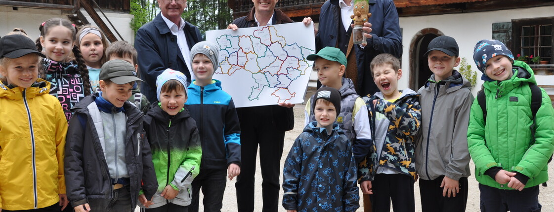 Caritas Direktor Johannes Dines  und der Salzburger Freichlichtmuseums-Direktor stehen mit Kindern auf einem Schotterplatz und halten ein Plakat hoch.
